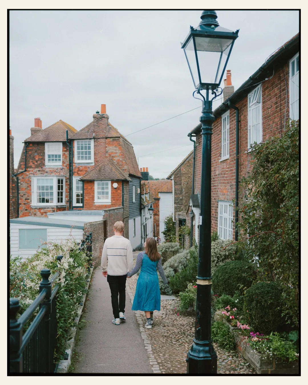 american couple walking down narrow path past a victorian street lamp in the ancient town of rye for their engagement shoot ahead of their destination wedding