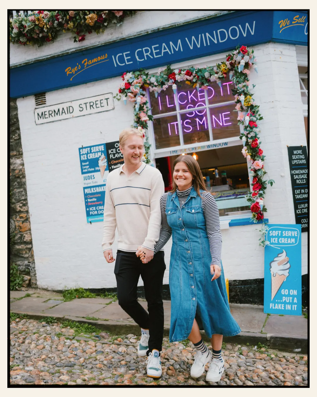 american couple walking in front of ice cream shop with neon sign i licked it so its mine on a cobbled narrow street in the ancient town of rye for their engagement shoot ahead of their destination wedding