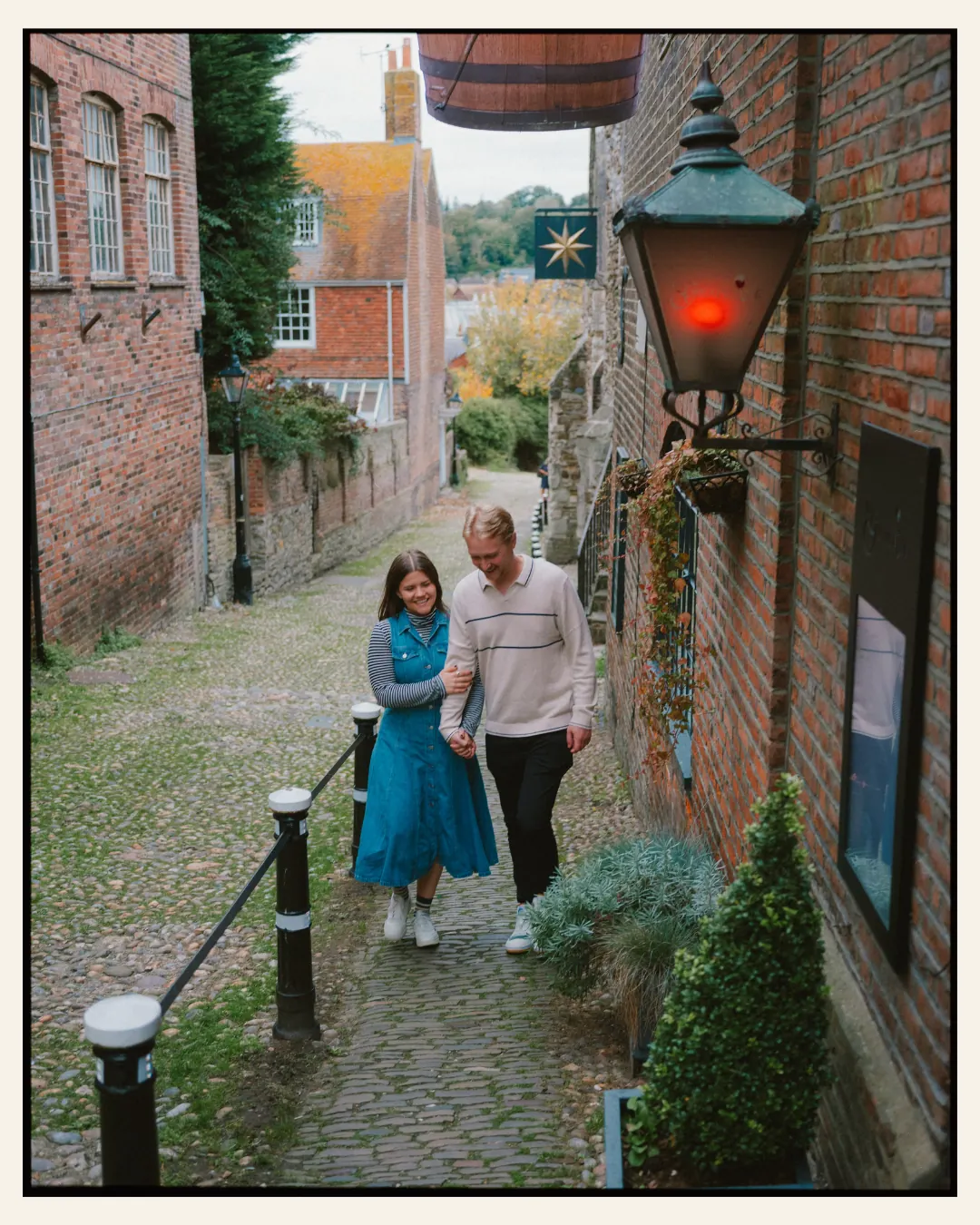 american couple walking up a cobbled narrow street with lantern in the ancient town of rye for their engagement shoot ahead of their destination wedding