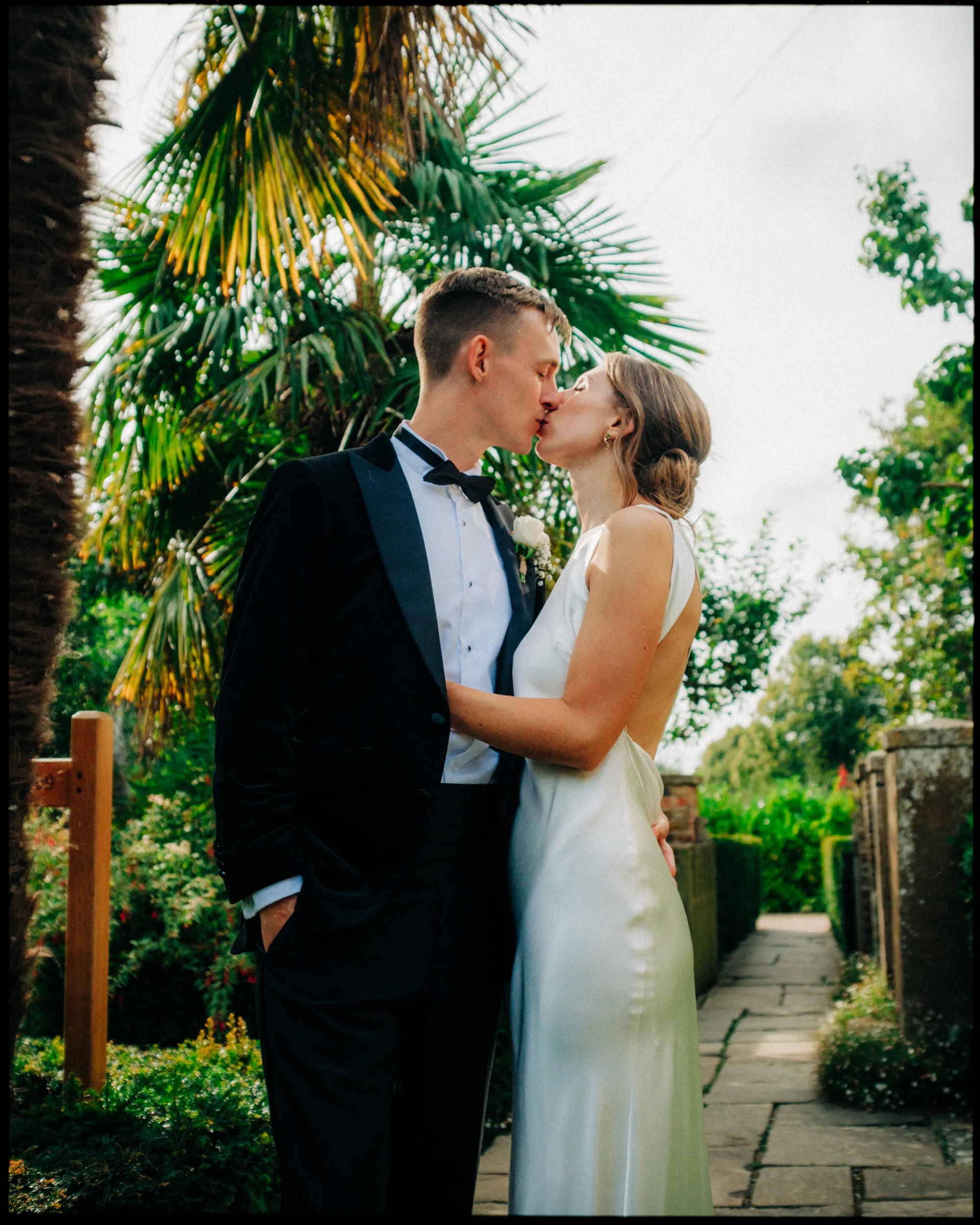 A bride in a white satin dress and a groom in a black tuxedo share a kiss outdoors on a stone path, surrounded by lush greenery and palm trees.