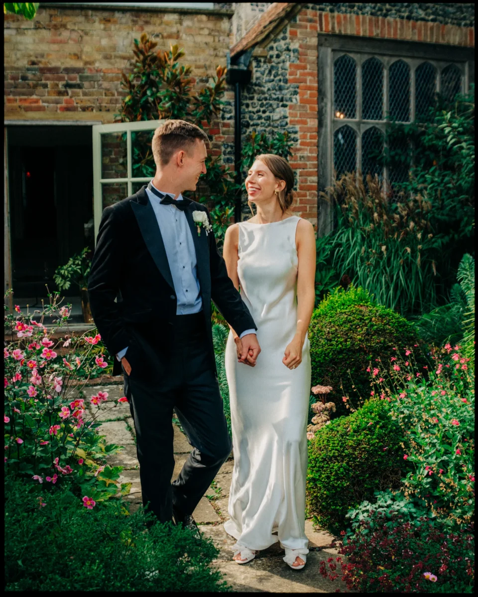 A couple in wedding attire, holding hands and smiling at each other, stands in a lush garden with blooming flowers and greenery, in front of a rustic brick building with large windows.