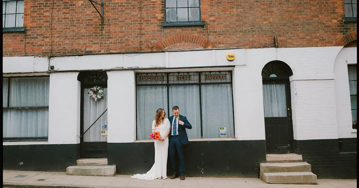 A bride in a white dress holding bright flowers stands next to a groom in a blue suit on a sidewalk in front of a brick building with large windows and a flower wreath on the left door.
