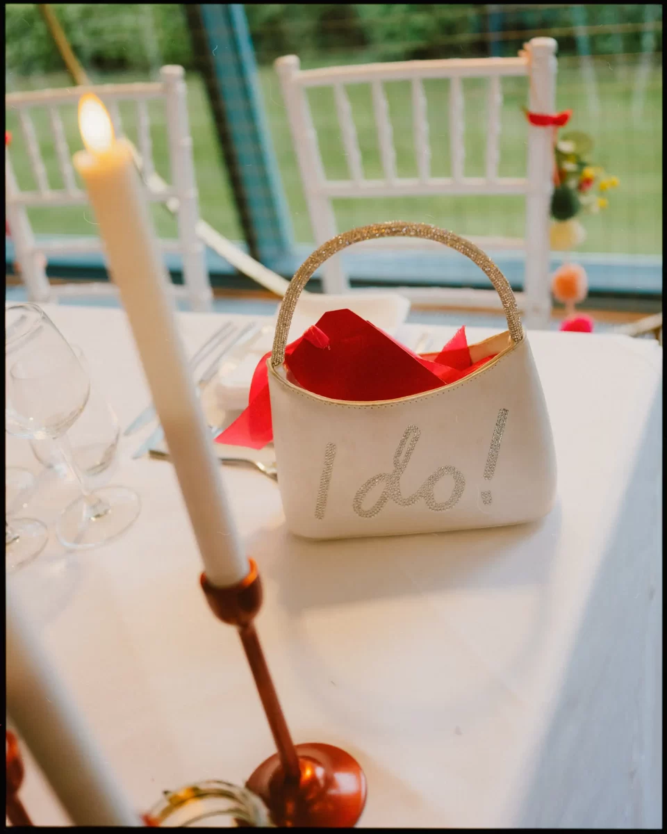 A white handbag with I do! written in glitter sits on a decorated table with red napkins, candles, wine glasses, and white chairs, suggesting a wedding celebration.