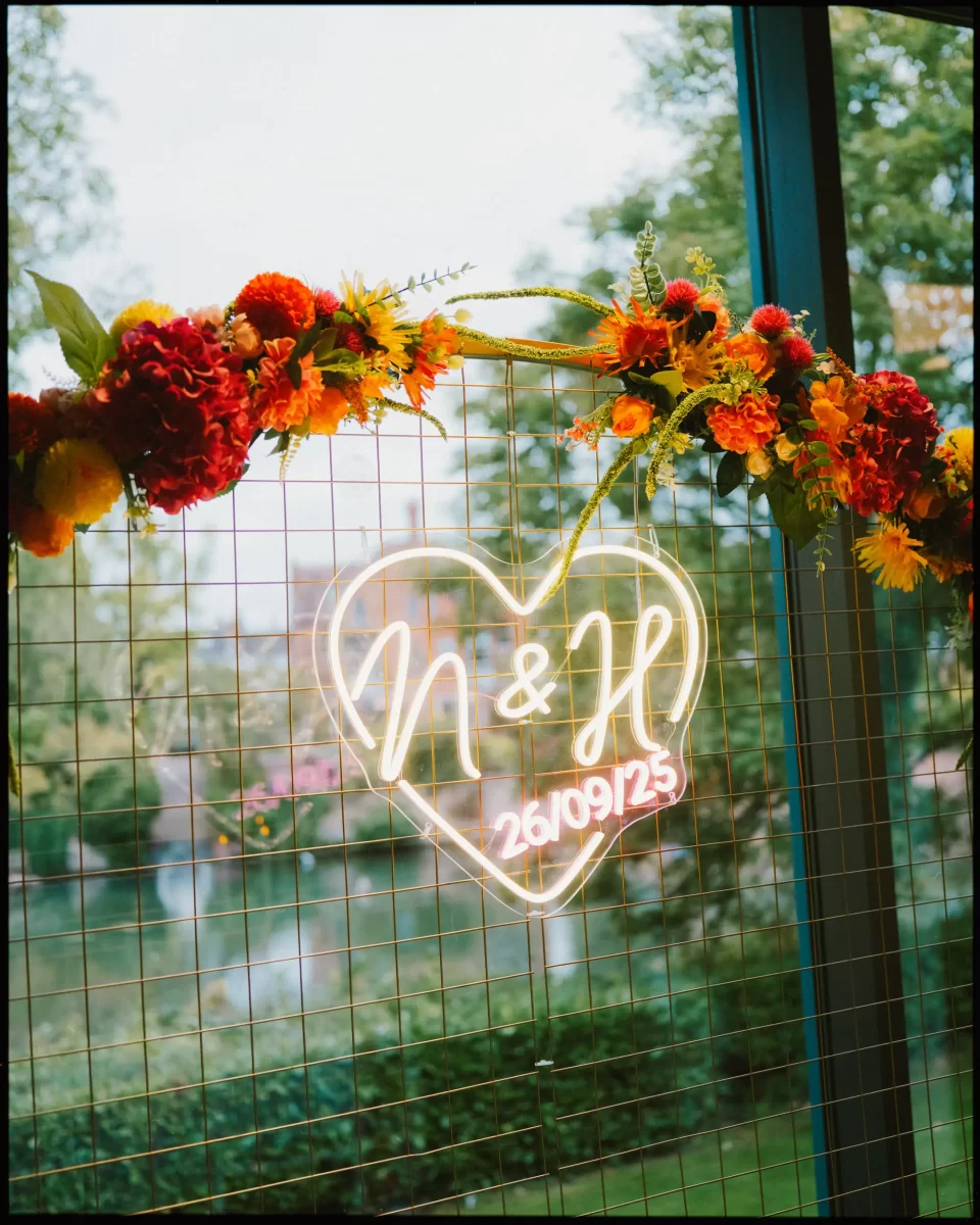 A neon heart sign with “N & H 26/09/25” hangs on a wire grid. The grid is decorated with a garland of red, orange, and yellow flowers. Trees and a pond are visible through the window behind it.