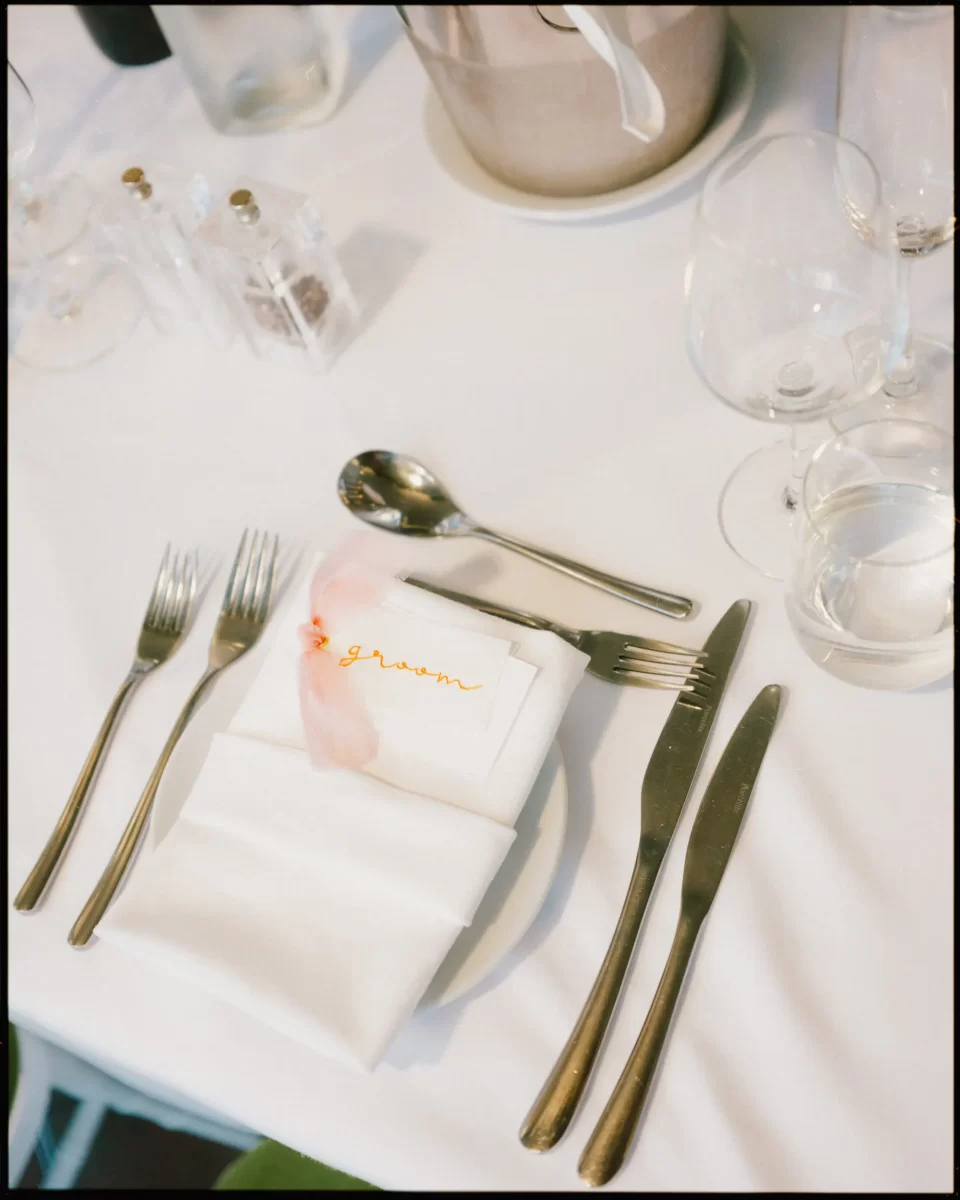 A white table setting with silver cutlery, wine glasses, and a napkin on a plate. A name card labeled groom with a pink ribbon rests on the napkin. Salt and pepper shakers and an ice bucket are in the background.
