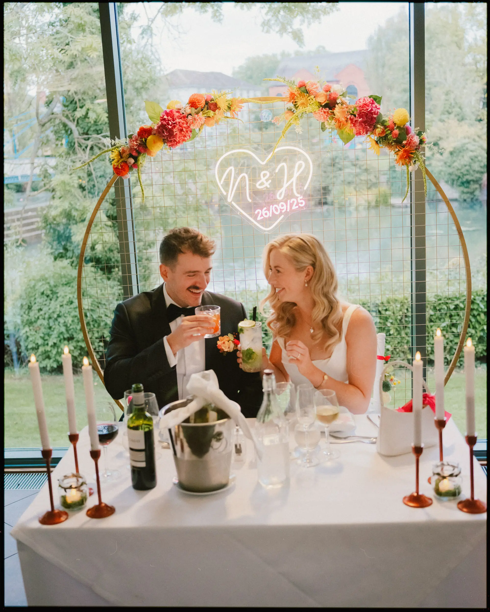 A bride and groom sit smiling at a decorated table, toasting drinks. Behind them is a floral arch with a neon heart sign displaying “N & H 26/09/23.” The background shows green trees and a river through large windows.