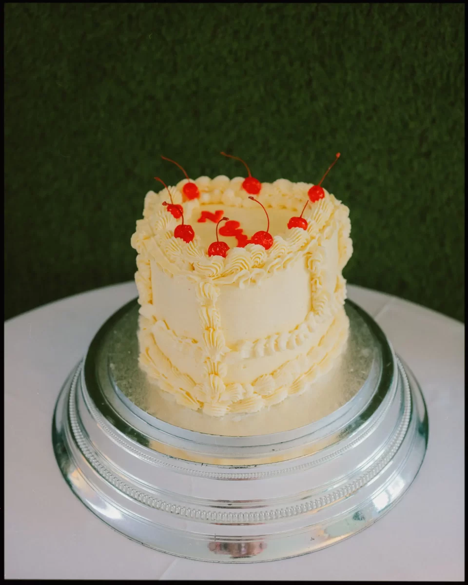 A round cake with yellow frosting and decorative piping is topped with several bright red cherries. The cake sits on a silver stand against a green textured background.