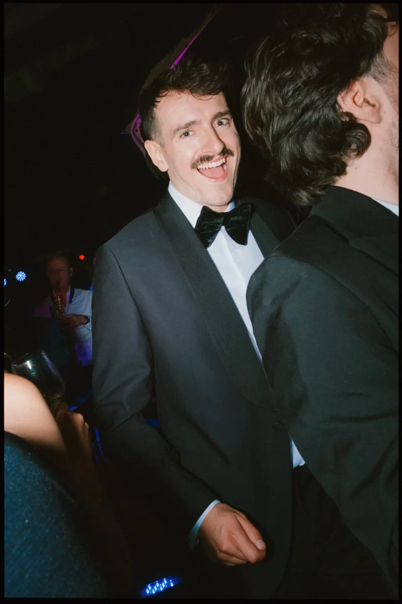 A man in a tuxedo with a bow tie smiles and looks at the camera while standing at a lively event. Another person in formal wear is in the foreground, partially out of frame. Musicians play in the background.