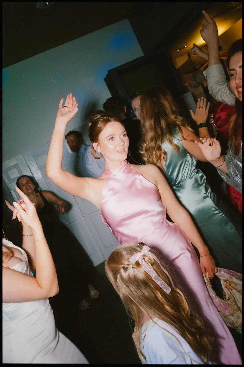 A woman in a shiny pink dress dances at a lively indoor party, surrounded by other people in formal attire, some smiling and raising their arms; a young girl with a ribbon in her hair stands nearby.