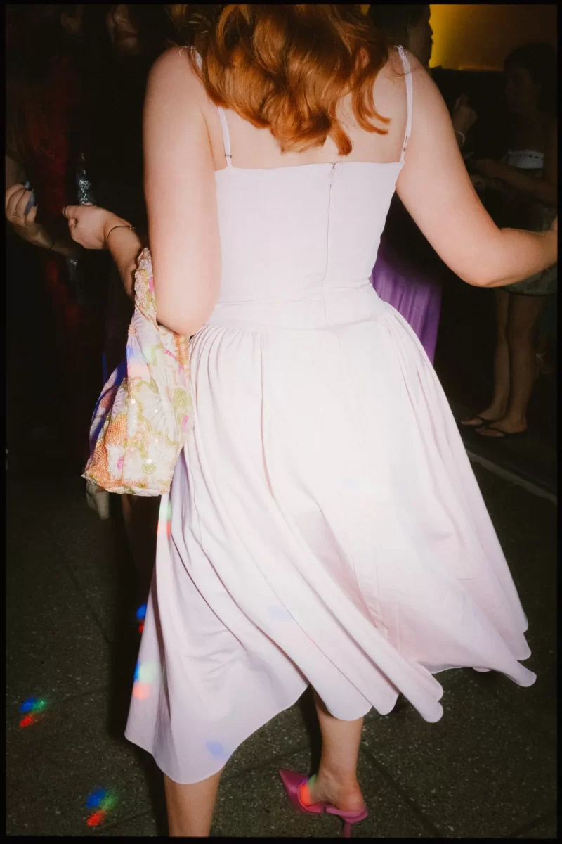 A person with red hair in a light pink dress and matching pink shoes dances at a party, holding a floral bag. Colorful lights are reflected on the ground and dress. Other people are visible in the background.