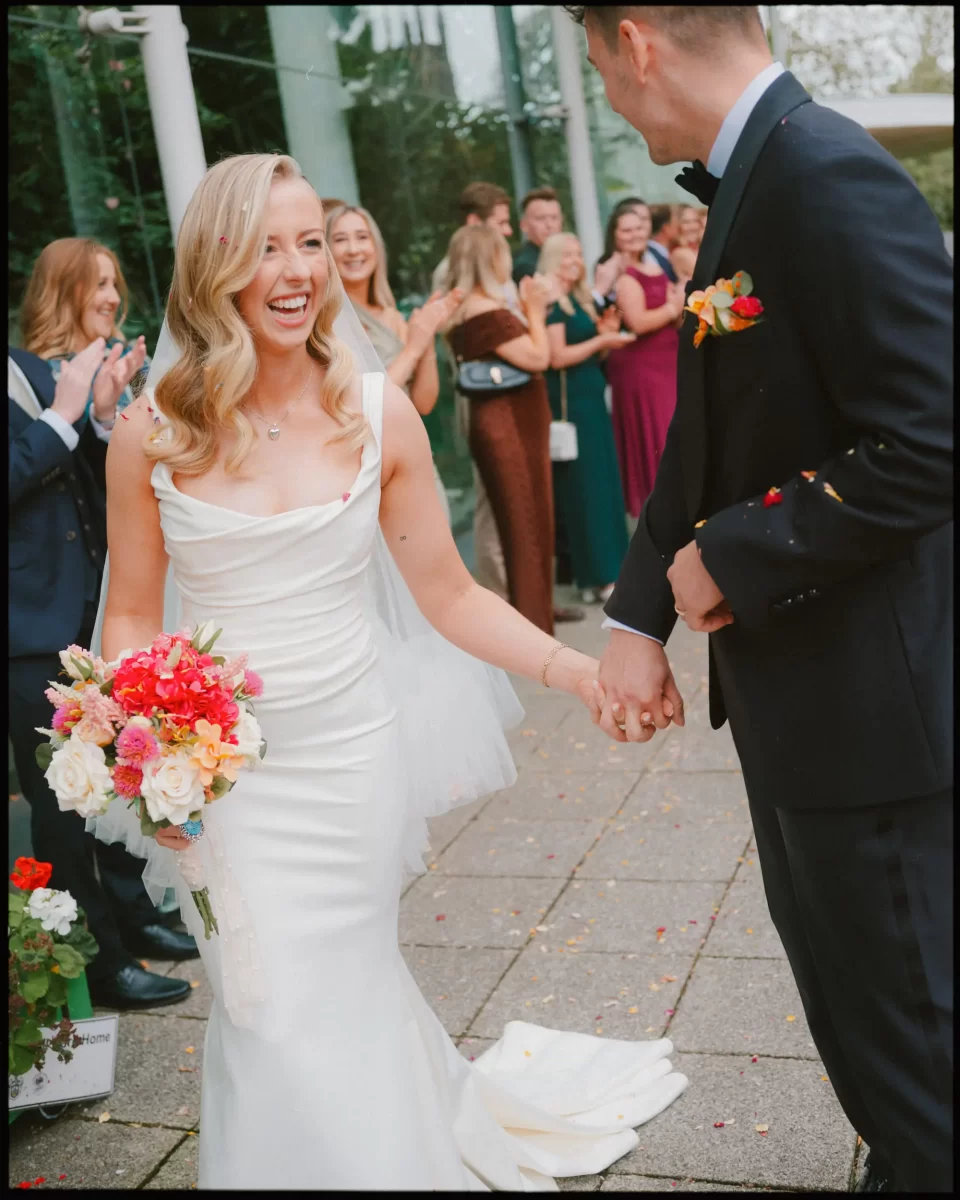 A smiling bride in a white dress holding a bouquet and the hand of a groom in a black suit, as wedding guests clap and cheer in the background outside a glass building.
