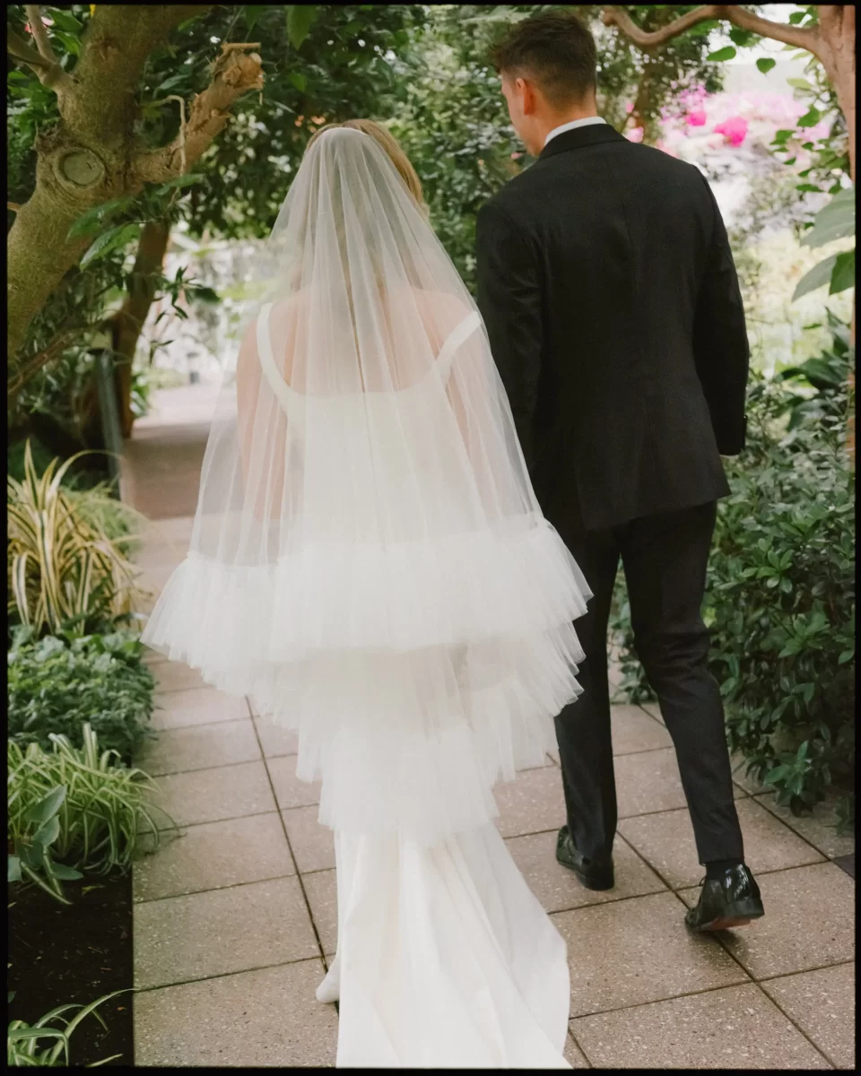 A bride in a white dress and veil walks beside a groom in a black suit along a garden path surrounded by lush greenery.