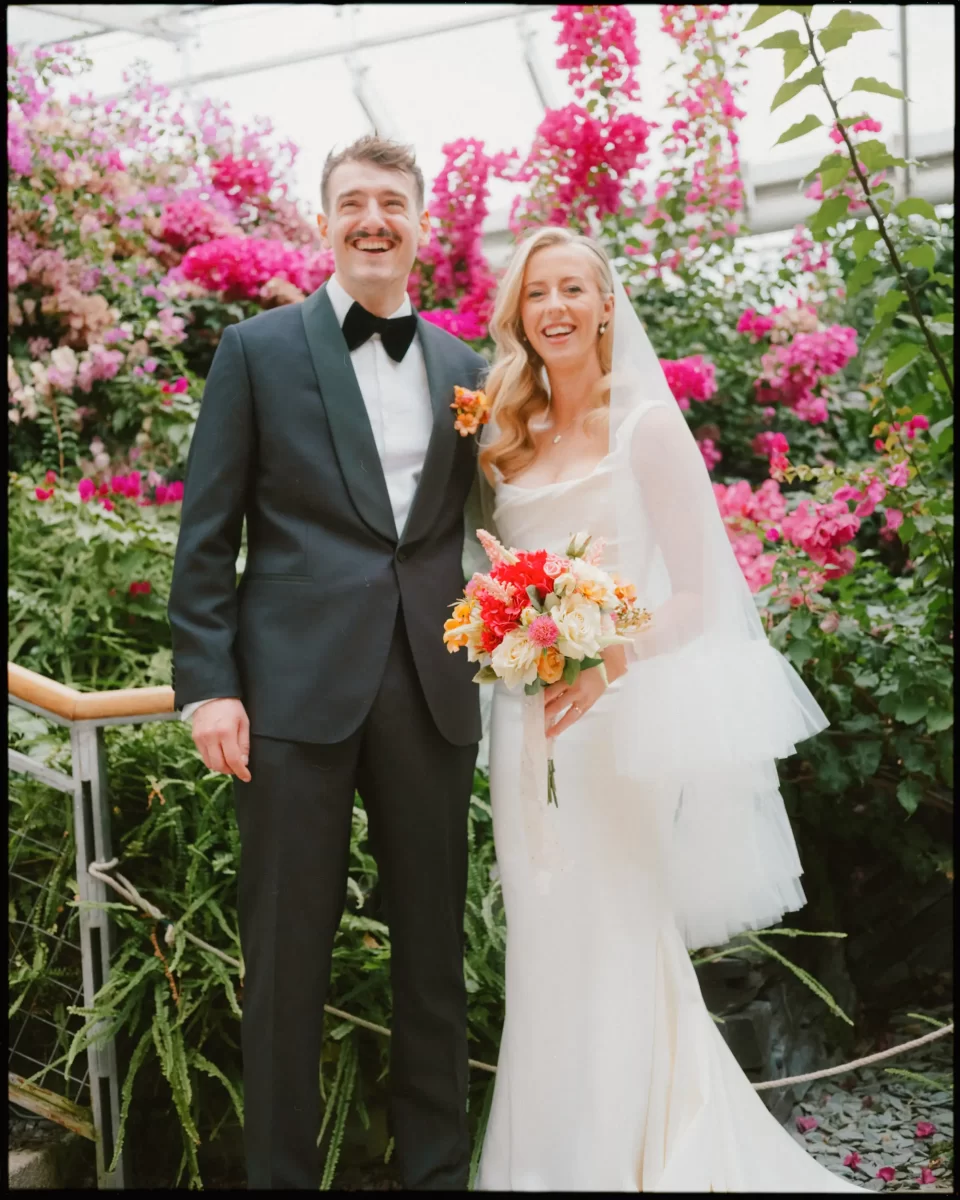 A bride in a white dress and veil holds a colorful bouquet while standing next to a groom in a black tuxedo. They smile in front of vibrant pink flowers and greenery.