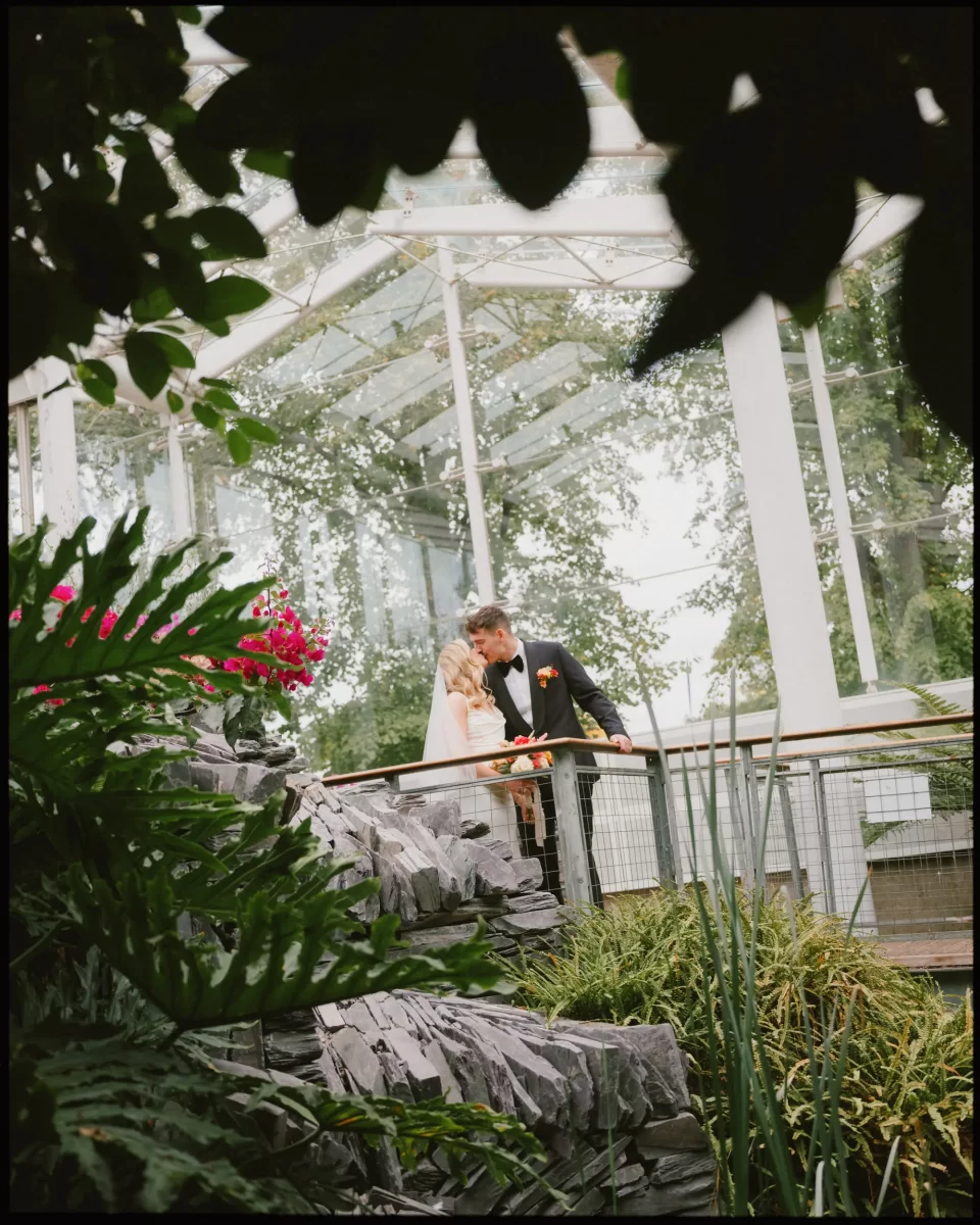 A bride and groom stand together on a wooden walkway surrounded by lush greenery and flowers, sharing a quiet moment under a glass canopy.