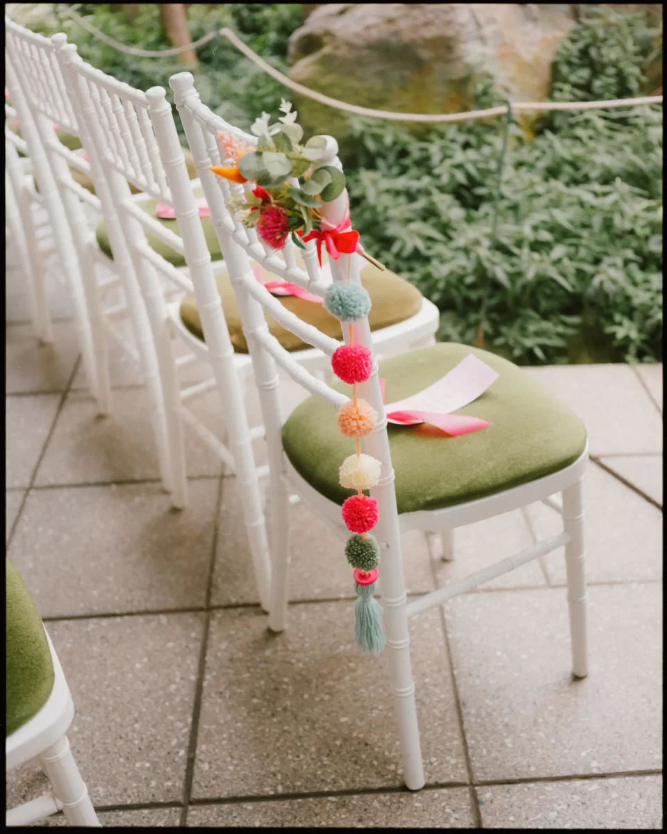White chairs with green cushions are decorated with colorful yarn pom-poms, pink ribbons, and greenery, set up outdoors on tiled flooring, surrounded by lush plants.