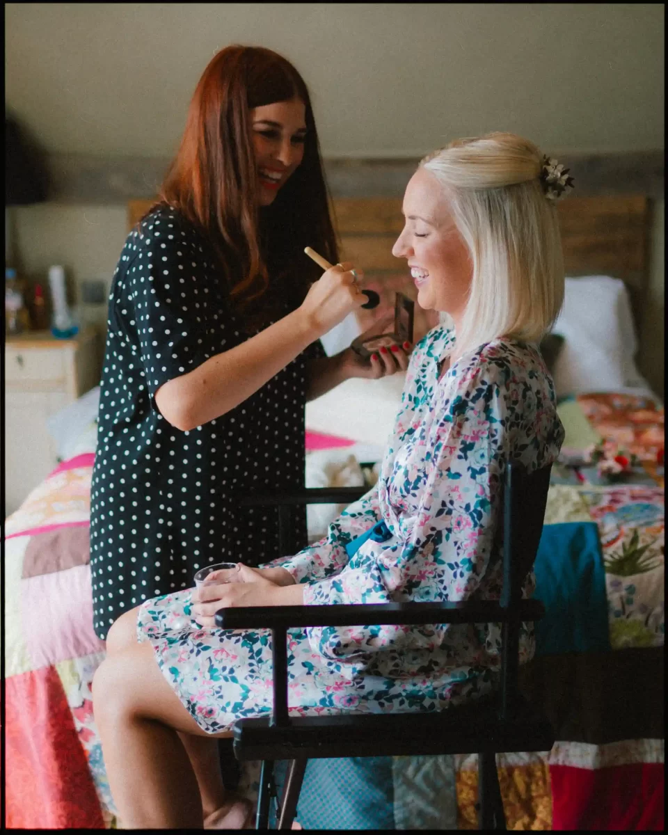 A woman with long brown hair applies makeup to a smiling blonde woman seated on a chair, both indoors near a bed with a colorful quilt. The blonde woman wears a floral robe.