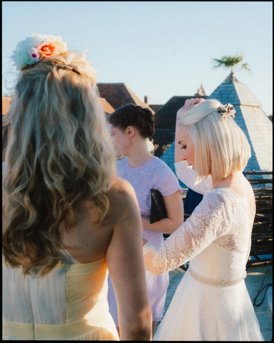 Three women wearing formal dresses stand outdoors in sunlight. One woman in a white lace dress adjusts her hair. Another, with long blonde hair and a floral headpiece, is partially turned away. Rooftops are visible in the background.