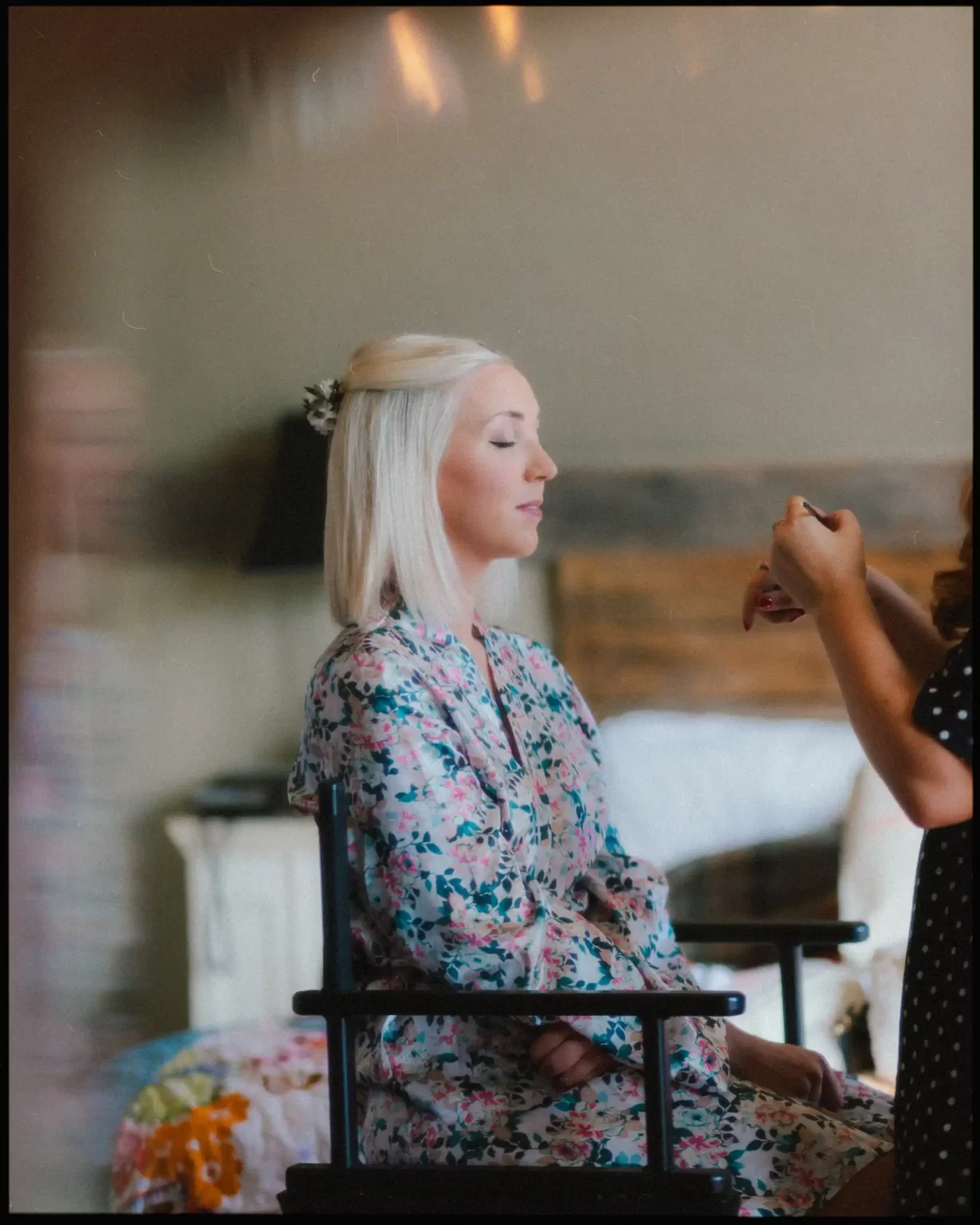 A woman with blonde hair, wearing a floral robe, sits with her eyes closed as another person applies makeup to her face in a softly lit room.