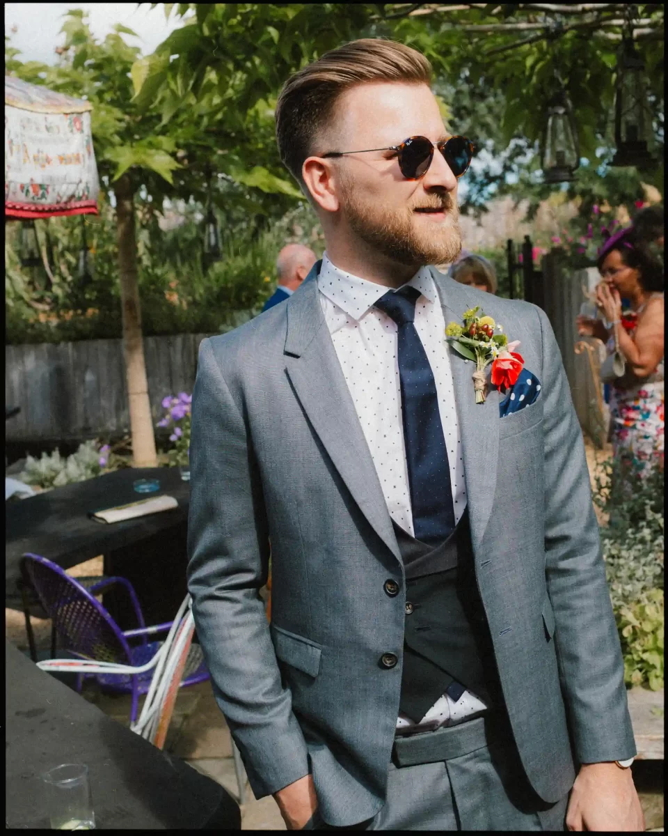 A man in a gray suit, polka dot shirt, dark tie, and sunglasses stands outdoors at a sunny event, wearing a colorful boutonniere, with people and greenery in the background.