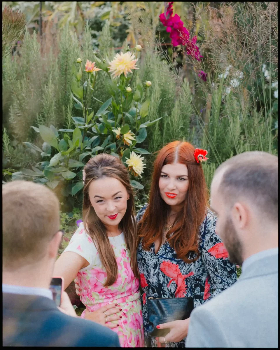 Two women pose and smile for a photo outdoors among lush flowering plants. Both wear bright, patterned dresses and have red lipstick. Two men stand in the foreground, one taking the picture with a phone.