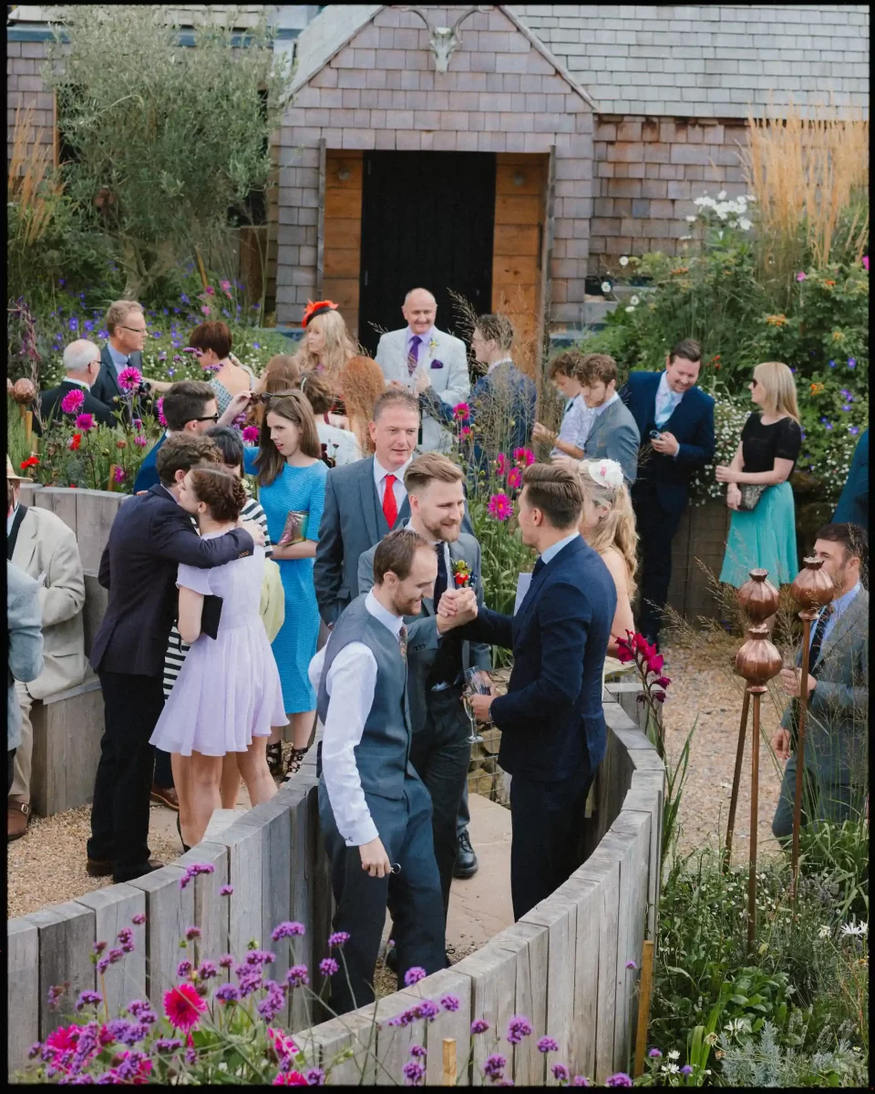 A group of people dressed in formal attire gather outdoors in a lush garden, chatting and celebrating near a rustic wooden building decorated with flowers and greenery.