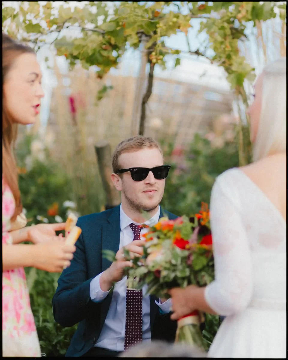 A man in a suit and sunglasses sits outdoors, holding something in his hands, while two women in dresses, one holding a bouquet of flowers, stand nearby. The background is filled with greenery and flowers.