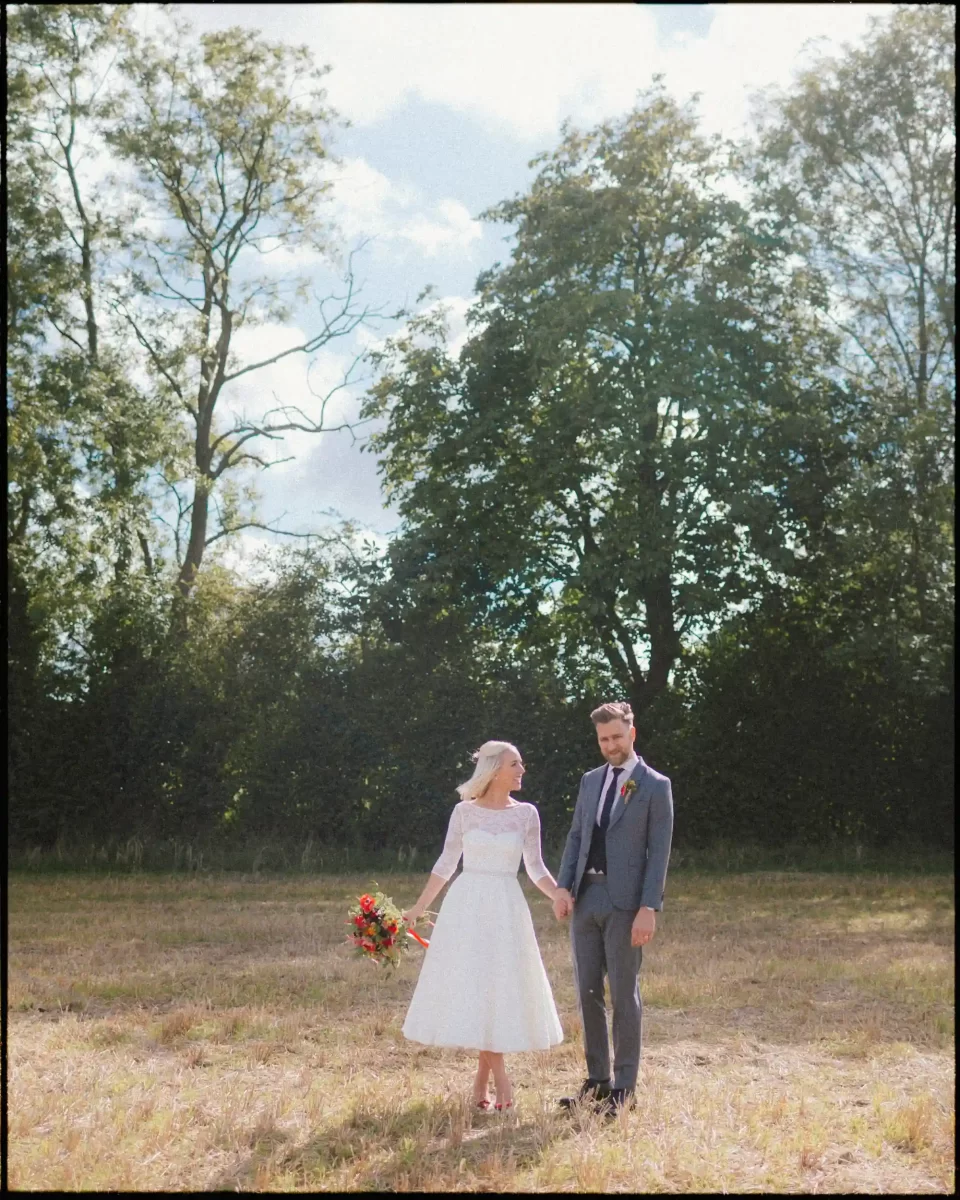 A bride in a white dress holding a bouquet and a groom in a gray suit stand together in a sunlit field, with tall green trees and blue sky in the background.