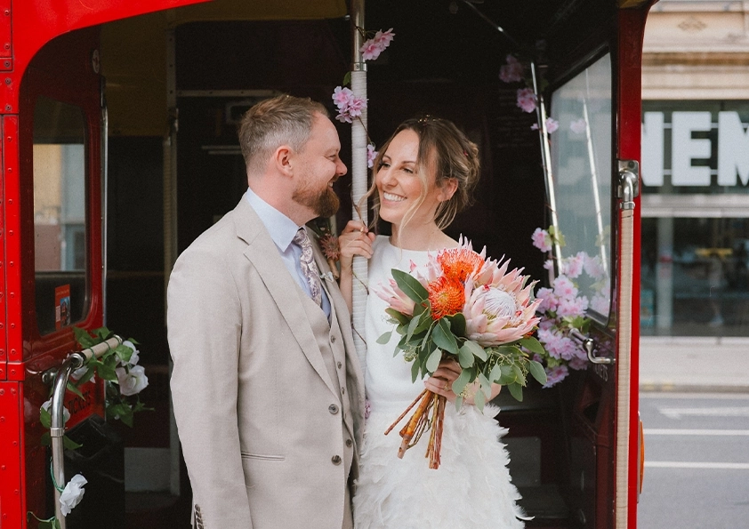 A newlywed couple smiles at each other in the doorway of a red bus at The Depot North London wedding venue. The bride holds a bouquet in her white dress, while the groom, in a beige suit, stands among pink floral decorations on the bus.