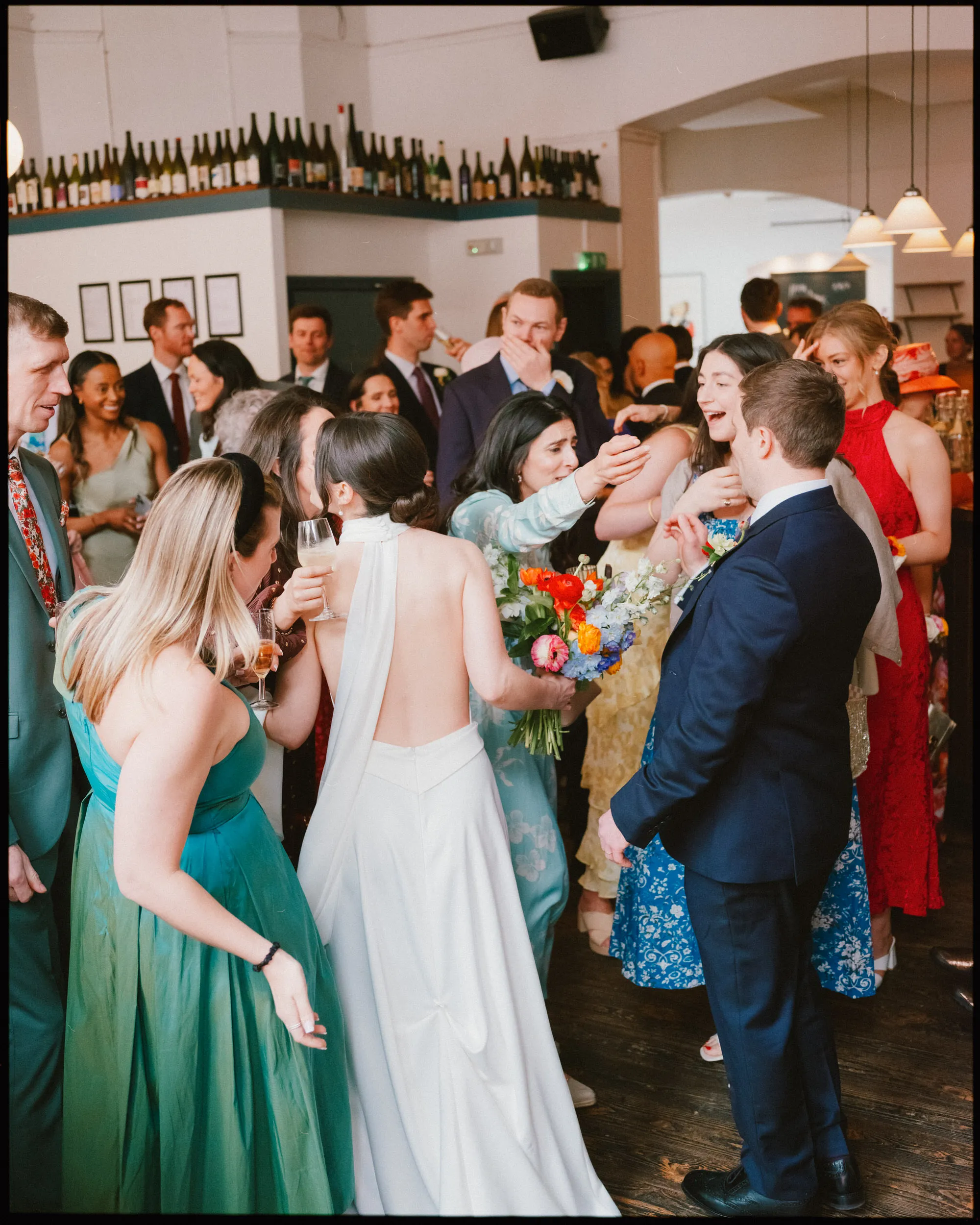 Bride in a backless white dress holds a bright bouquet as laughing guests with drinks crowd around her and the groom