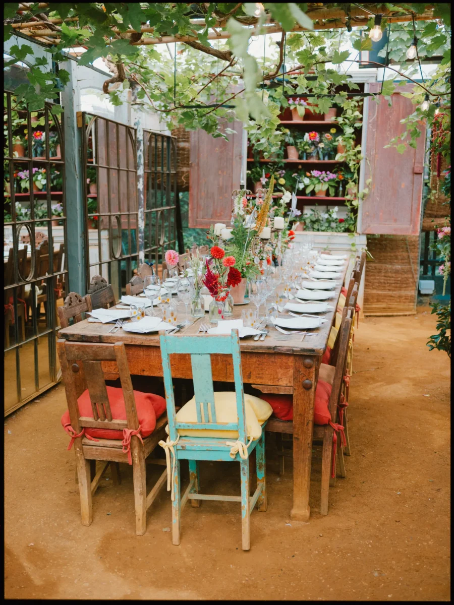 Long wooden dining table set with dahlias and candles under a vine-covered glasshouse roof at Petersham Nurseries