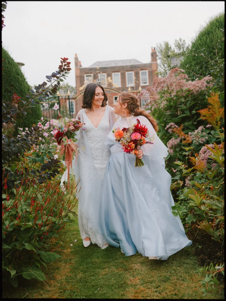 Two brides smile as they walk together through the colourful gardens at Petersham Nurseries, bouquets in hand