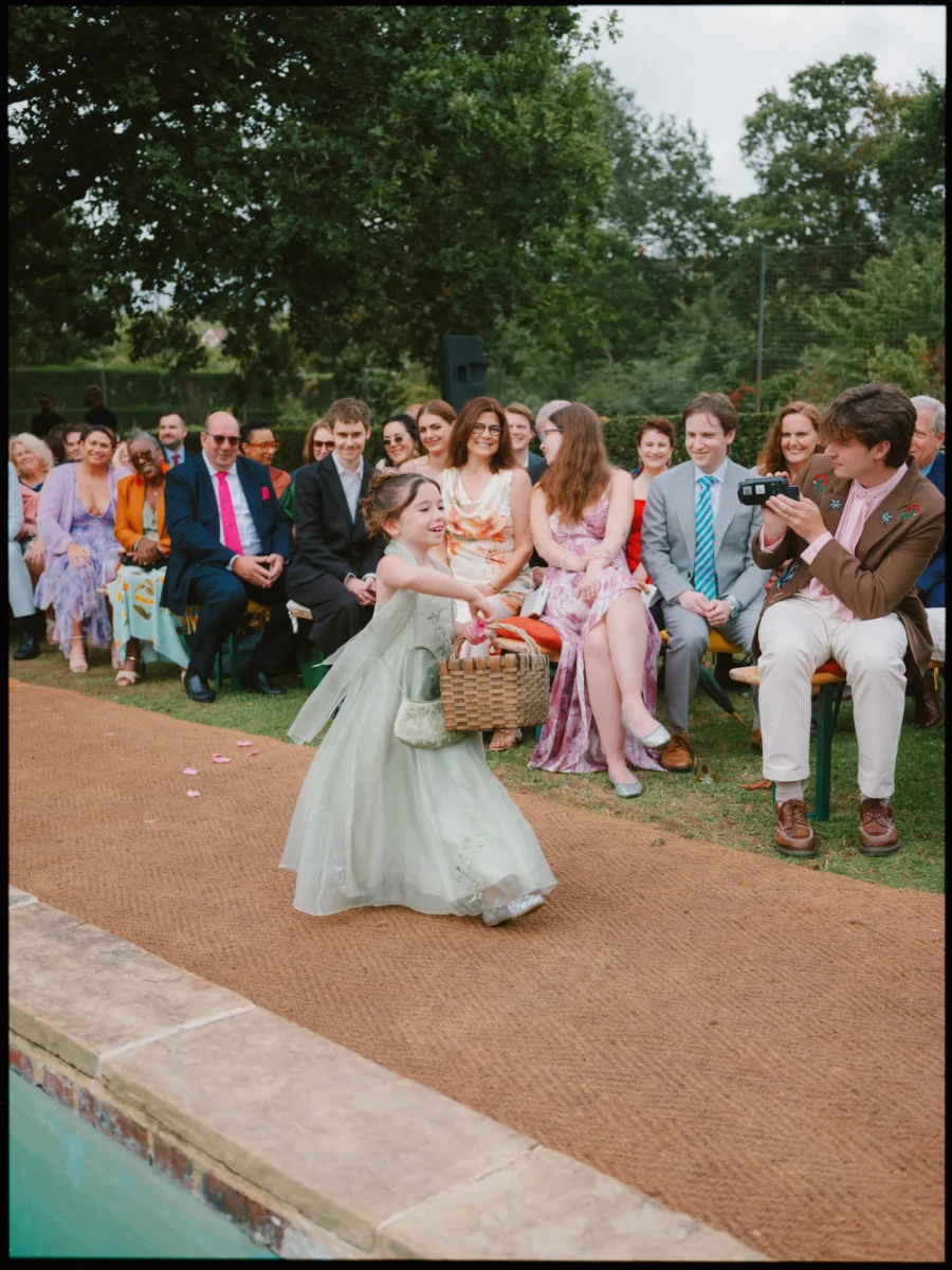 Flower girl in a pale green dress scatters petals down the coir aisle as smiling guests watch from the garden seats