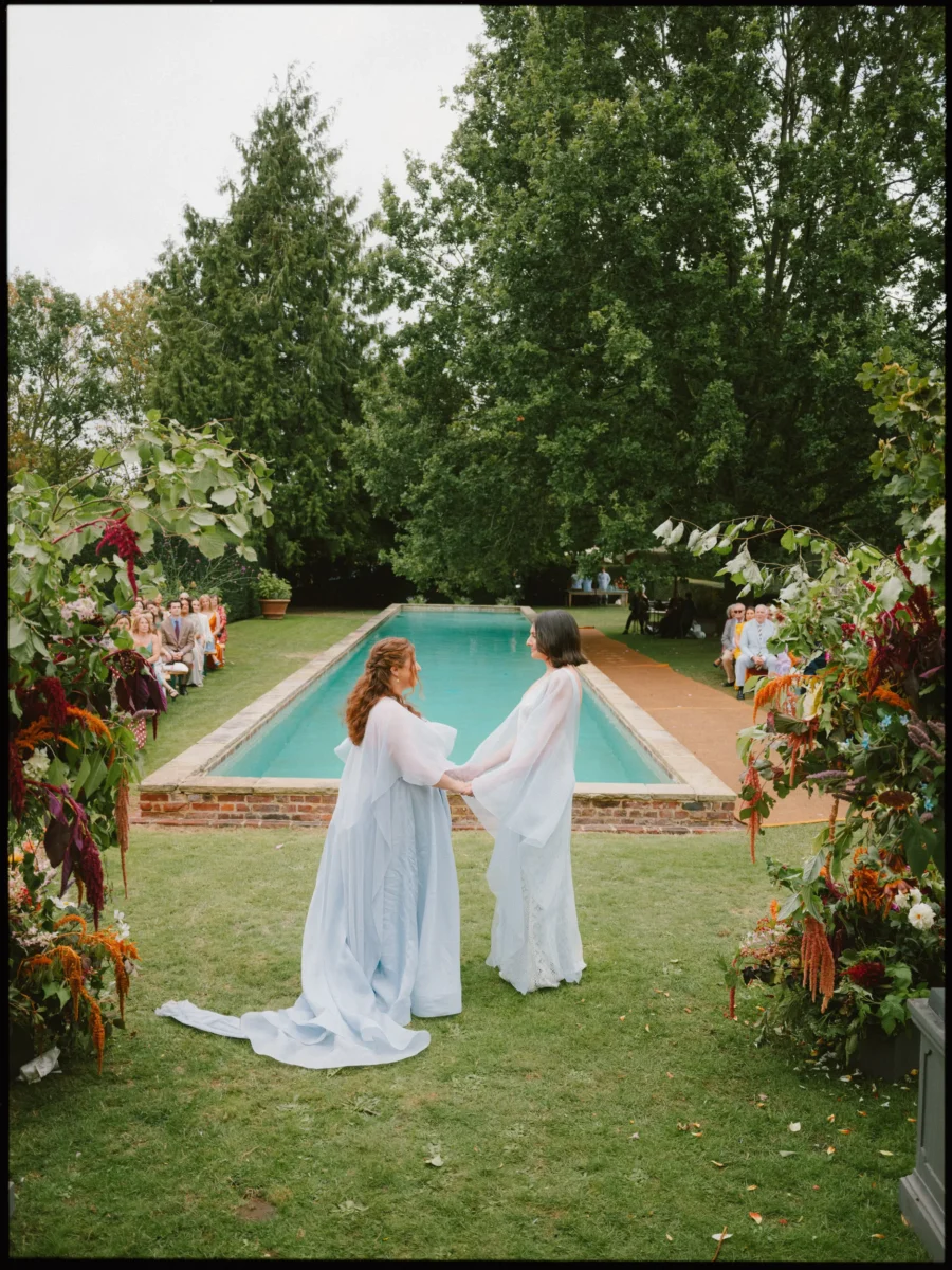 Two brides in pale blue and white hold hands beside a long pool as guests watch their outdoor ceremony at Petersham Nurseries