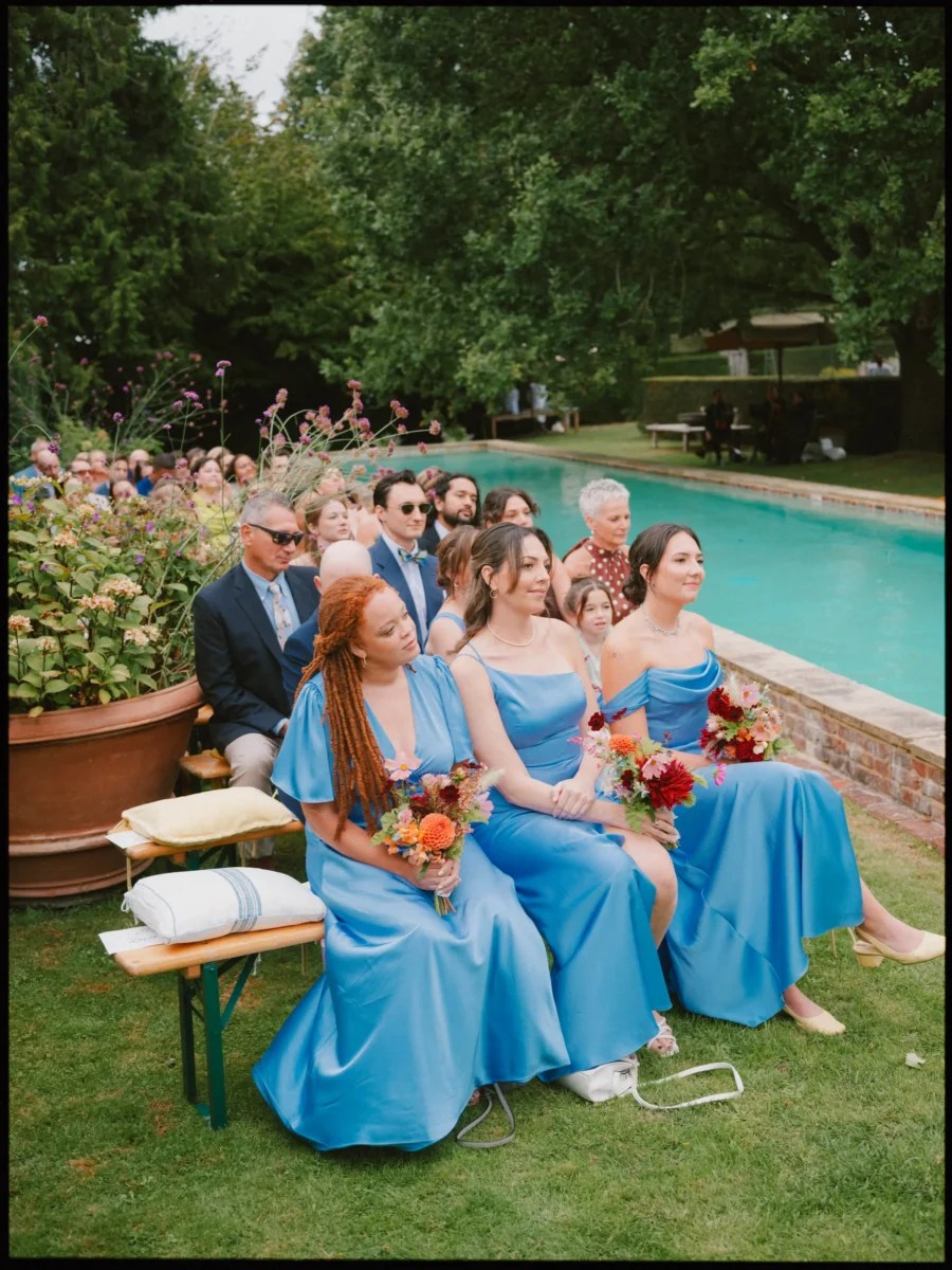 Bridesmaids in satin blue dresses hold bright bouquets on benches as guests watch the poolside ceremony
