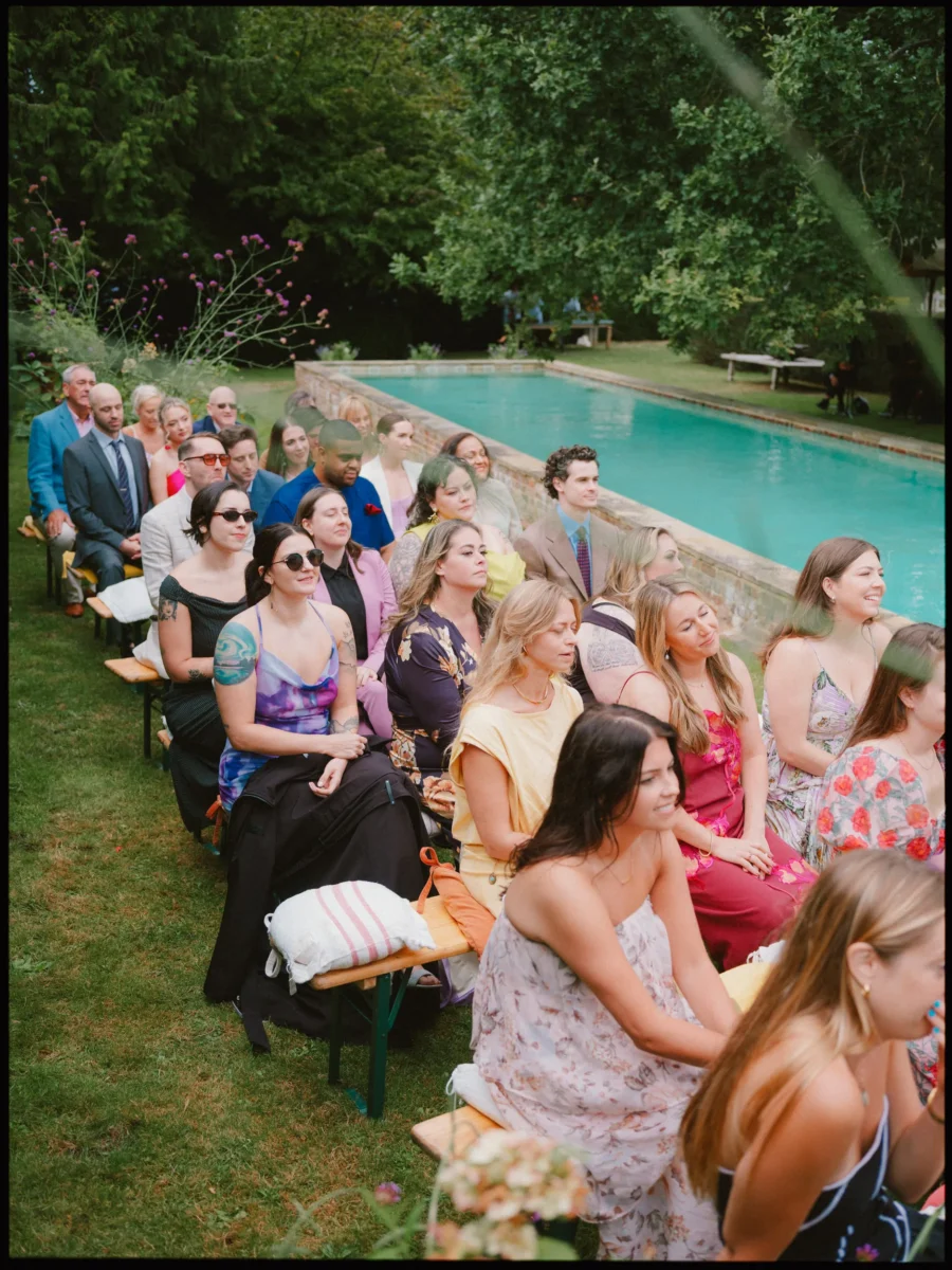 Smiling guests on wooden benches watch the ceremony beside the pool at Petersham Nurseries