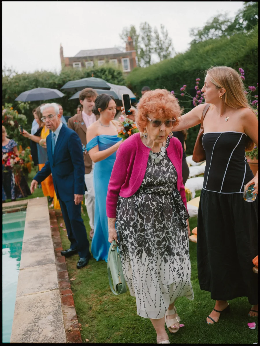 Older guest in a pink cardigan and sunglasses walks across the lawn beside the pool as other guests follow in the rain