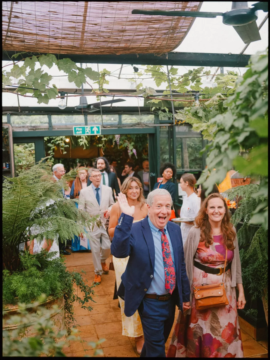 Guests stream into the Petersham Nurseries glasshouse, a man in a navy blazer waving and laughing among ferns and vines