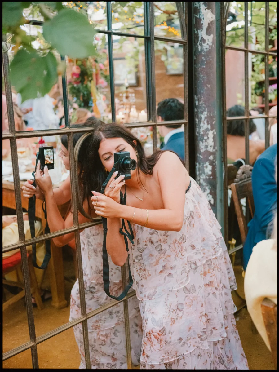 Guest in a floral ruffled dress snaps a photo through a metal-framed glasshouse window, her reflection doing the same