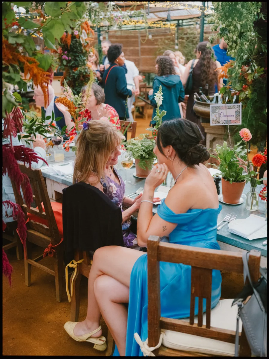 Two guests in a blue off-the-shoulder dress and floral top chat at a long table among foliage in the Petersham glasshouse