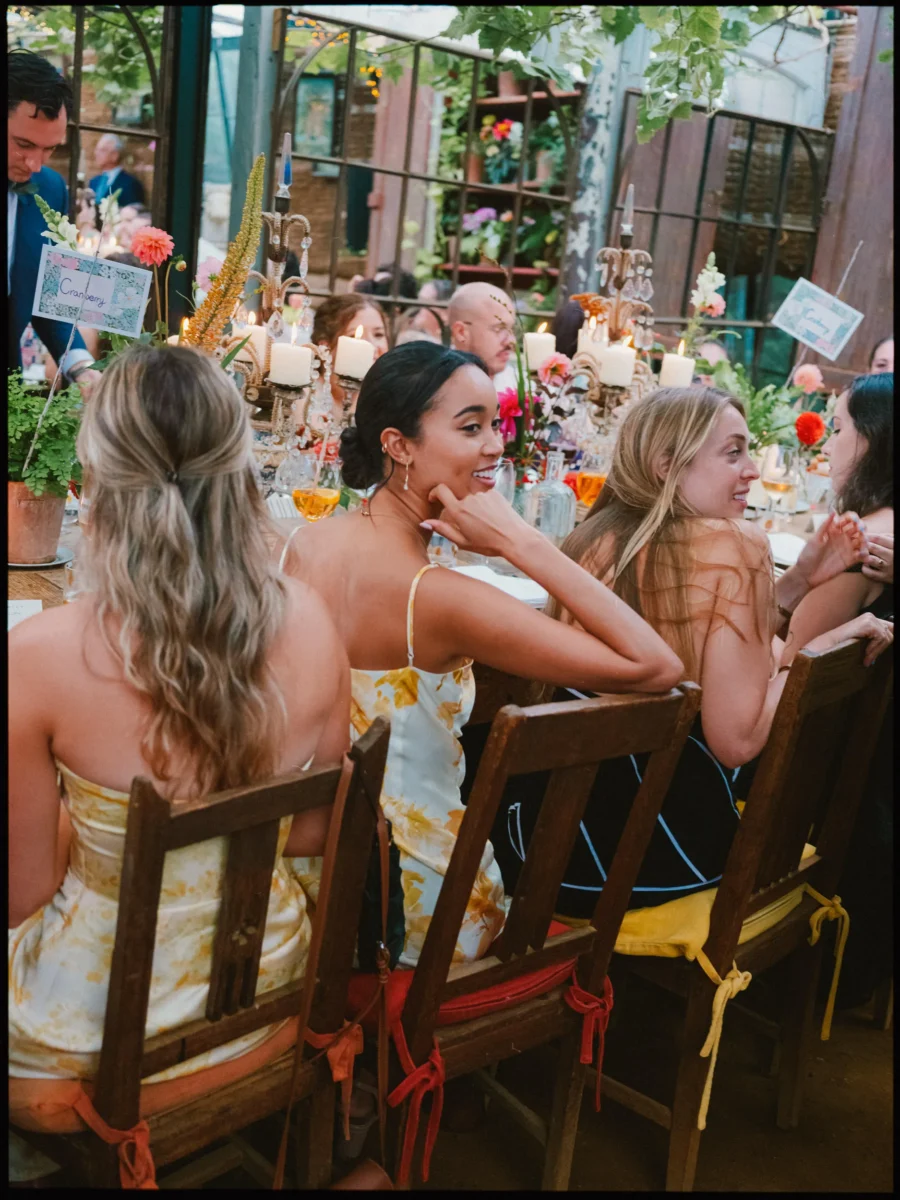 Guest in a yellow floral slip dress glances back with a smile at a candlelit dinner table in the Petersham glasshouse