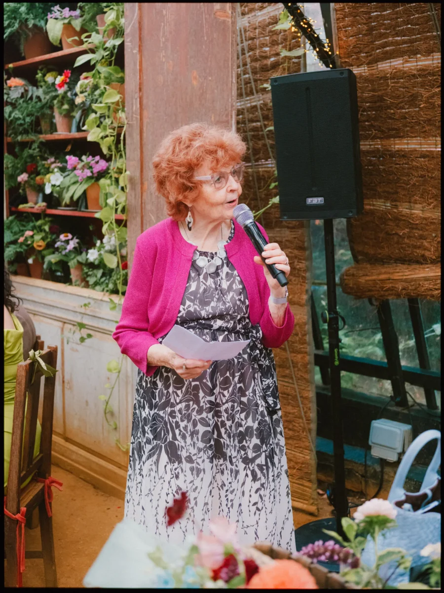 Older woman in a pink cardigan reads her notes into a microphone while giving a speech in the plant-filled glasshouse