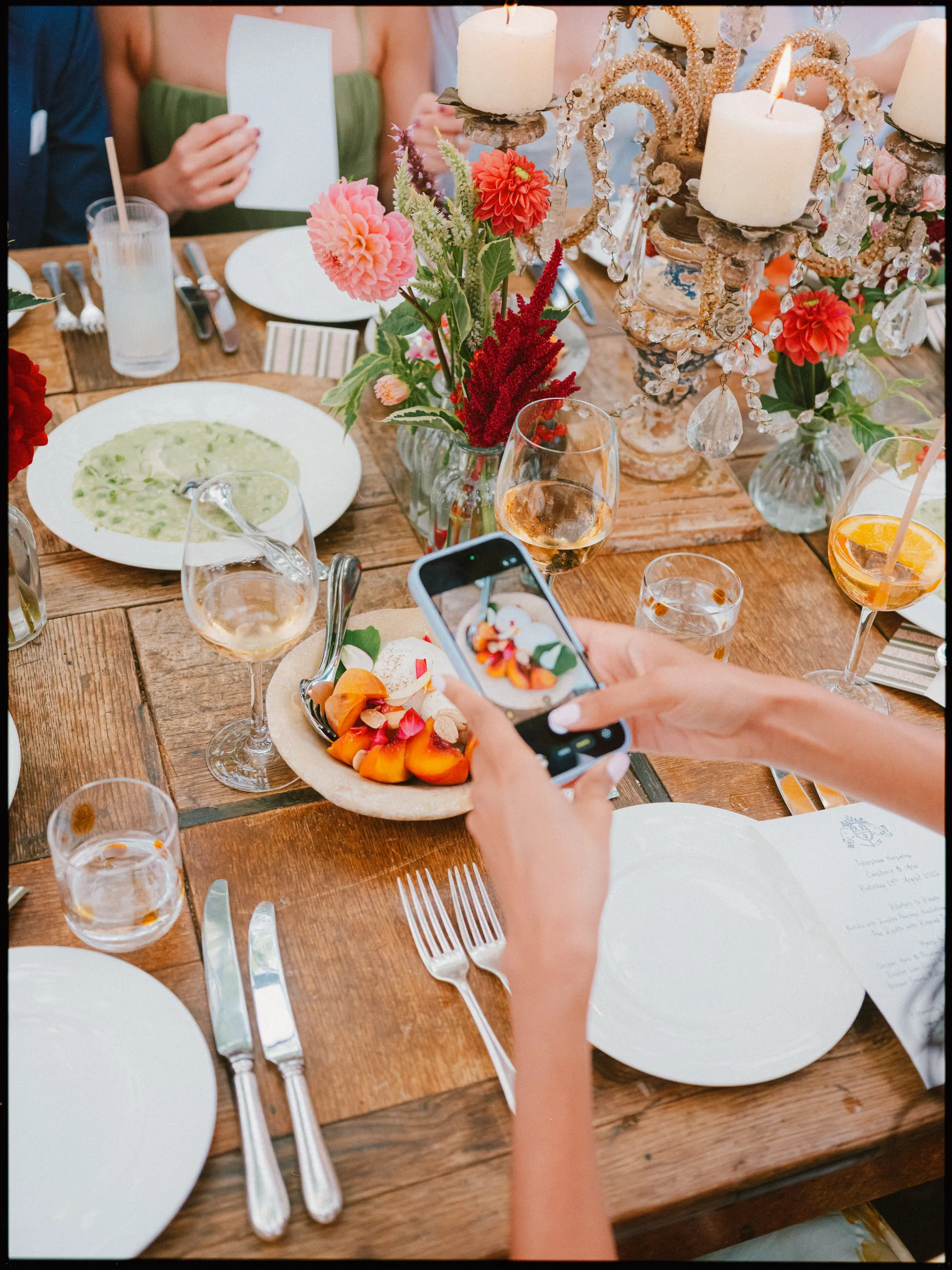 Guest photographs a plate of peaches and burrata on a rustic wedding table with dahlias and candles at Petersham Nurseries