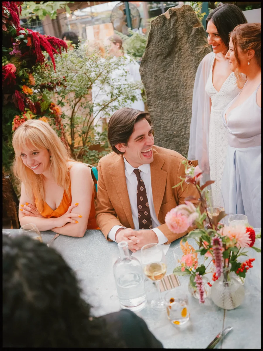 Guest in a tan jacket laughs by pink dahlias as two women in pale dresses smile at Petersham Nurseries
