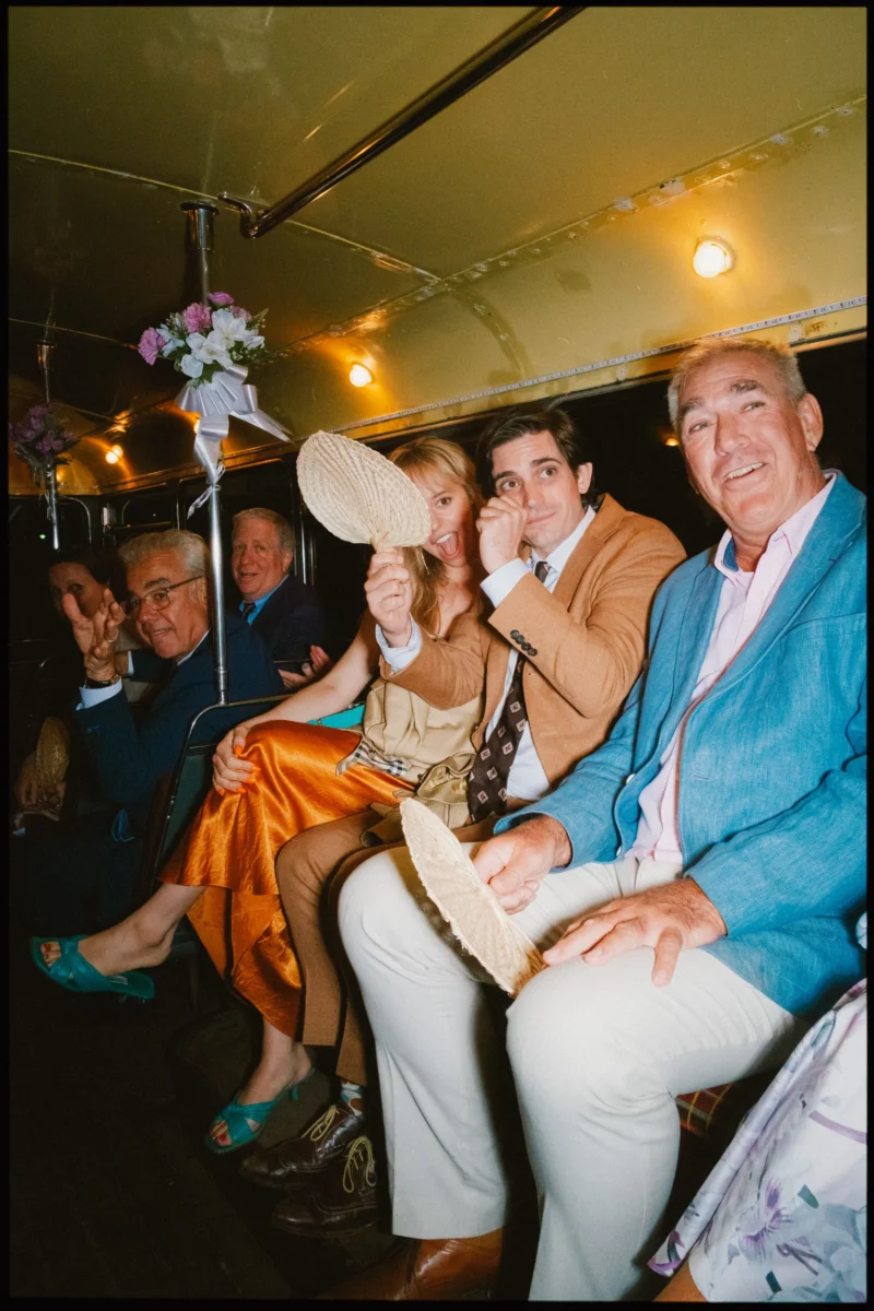 Guests grin and wave woven fans from the seats of the late-evening wedding bus, one pulling a delighted face