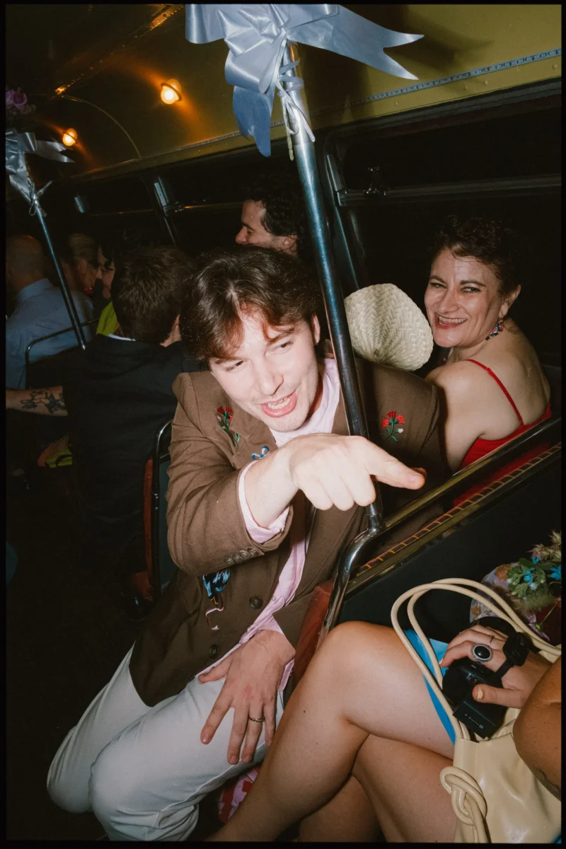 Guest in a brown jacket points at the camera from a packed wedding bus while a woman in red laughs behind him