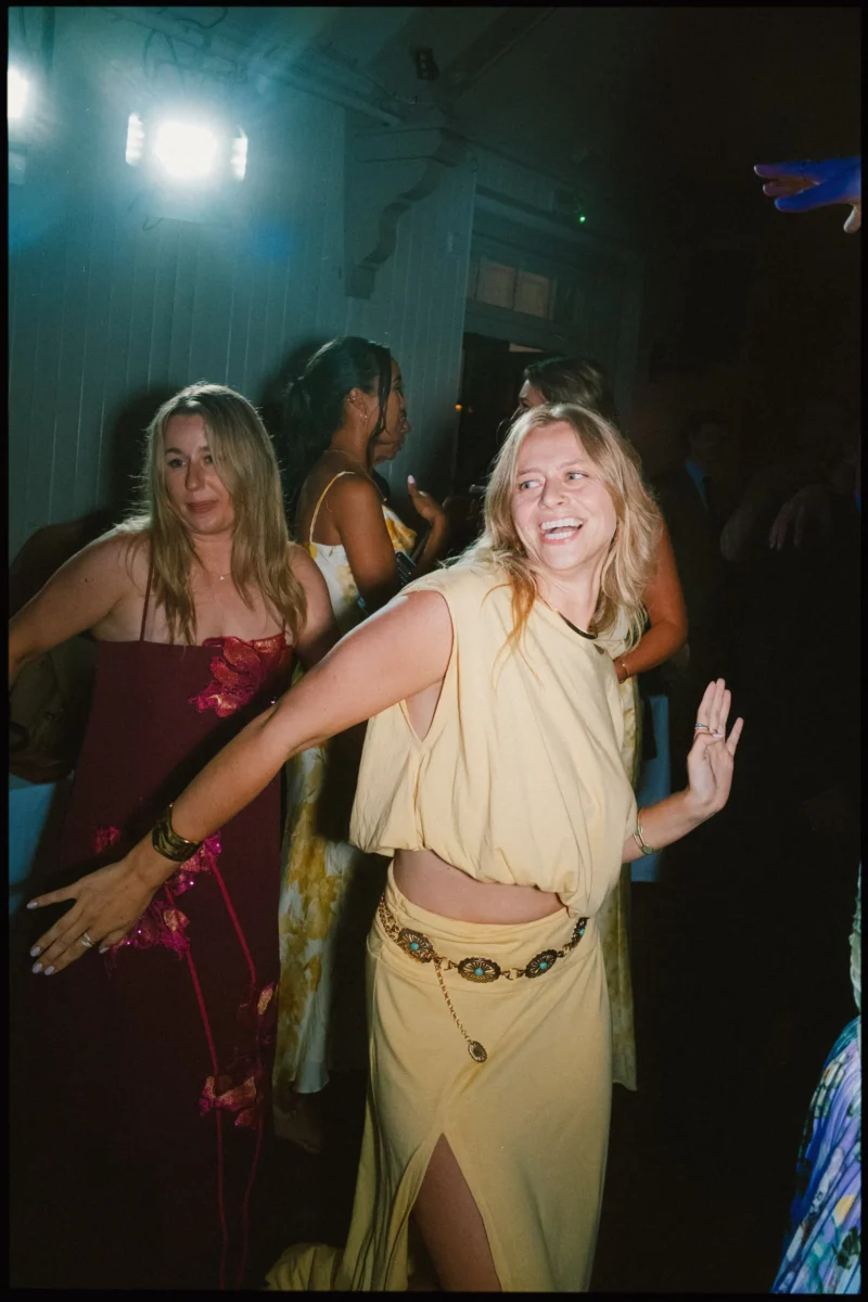 Woman in a pale yellow top and skirt beams mid-dance, arms outstretched, as guests dance behind her late at night