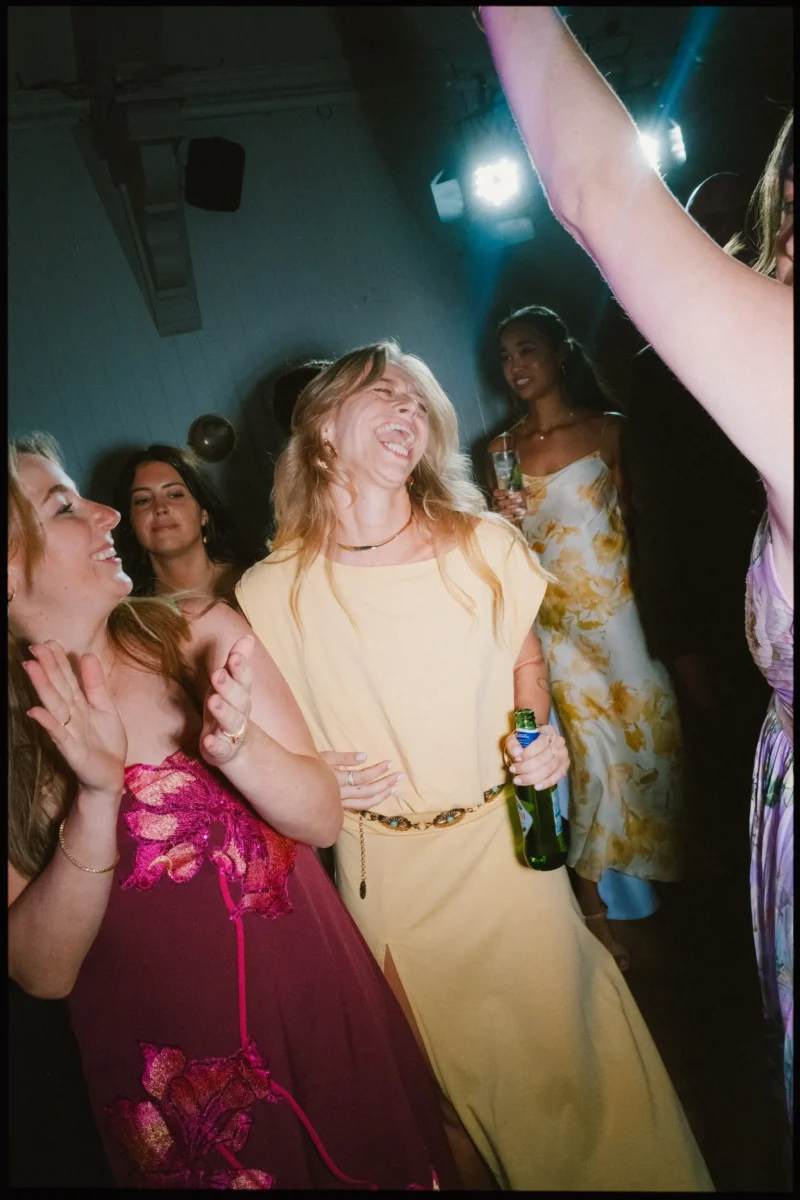 Woman in a yellow dress laughs with her head back, beer in hand, as guests dance and clap under party lights