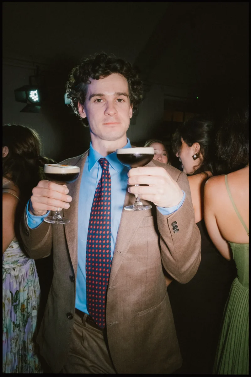Guest in a tan blazer and blue shirt holds up two espresso martinis to the camera on the late-night dance floor