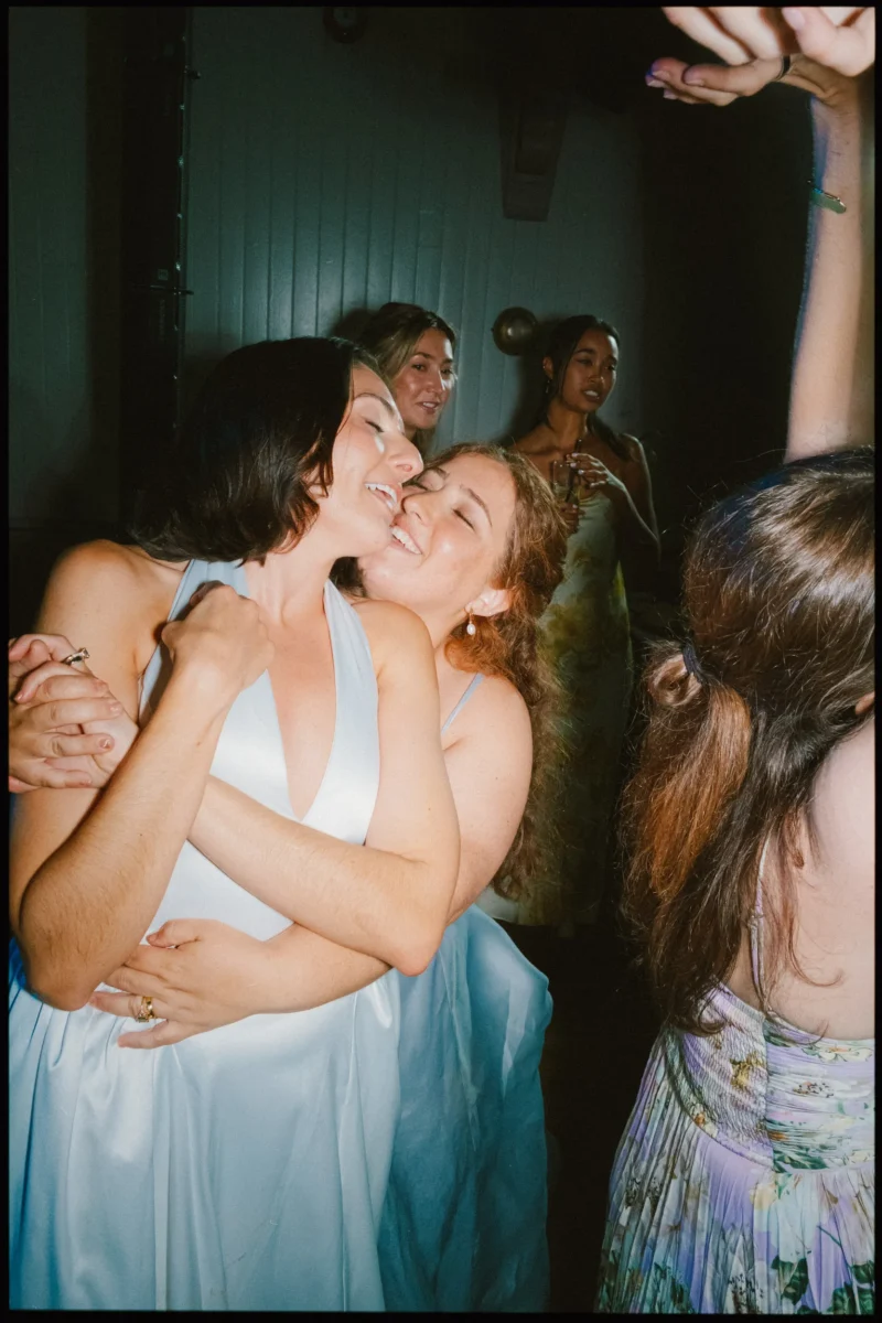 Two women in pale blue dresses hug cheek to cheek, laughing, as guests dance around them in a dimly lit room