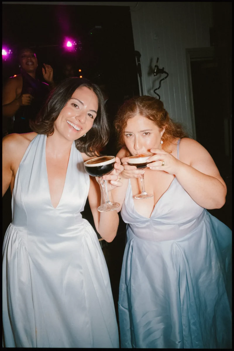 Two women in pale satin dresses hold espresso martinis at the late-night party, one grinning, one mid-sip